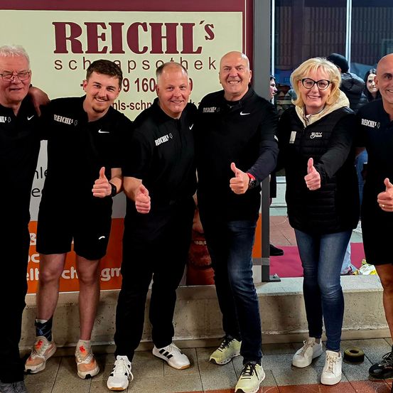 Sechs Menschen stehen in einer Reihe, lächeln und geben Daumen hoch vor einem Schild für Reichl's Schnapps.
