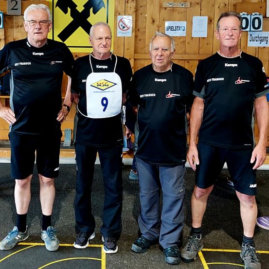 Vier Männer in schwarzen Trikots stehen zusammen in einem Fitnessstudio. Einer hat ein weißes Startnummernband mit der Nummer 9. Dahinter ein gelbes Schild mit einem X und eine blaue Nummer 2. Im Hintergrund eine hölzerne Wand mit verschiedenen Schildern und einem Regal.