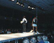 Zwei Personen stehen auf der Bühne, eine hält einen Ball und die andere ist in Tenniskleidung. Ein Publikum sitzt unten und schaut unter einer gedämpften Decke mit mehreren Lichtern.