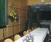 Ein Konferenzraum mit einem langen Tisch, weißer Tischdecke, Blumen, Stühlen und einer Leiter in der Nähe einer Pflanze. Der Raum hat Holzwände und einen grünen Vorhang.