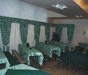 Ein leerer Raum mit mehreren Tischen und Stühlen, die mit grünen und weißen Tischdecken bedeckt sind. Die Wände sind mit grünen und weißen Vorhängen geschmückt, und es steht eine Pflanze in der Ecke.