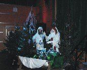 Zwei Personen in weißen Kostümen stehen auf einer Bühne, die mit Weihnachtsbäumen und einem weißen Tuch geschmückt ist. Die Umgebung erscheint schwach beleuchtet, mit einer hölzernen Wand und schwarzen Vorhängen im Hintergrund.