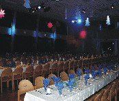 Ein großer Bankettsaal mit vielen Tischen für eine formelle Veranstaltung. Die Tische sind mit weißen Tüchern und blauen Servietten bedeckt, die in Form einer Blume gefaltet sind. Der Saal ist mit blauen und roten Lichtern und Weihnachtsdekorationen, die von der Decke hängen, geschmückt.