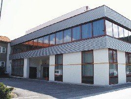 Ein modernes Gebäude mit Glasfenstern und einem grauen Dach, das sich in einem Parkplatz befindet. Das Gebäude ist weiß mit Glaspaneelen und einem großen Eingang.