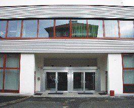 Ein modernes Gebäude mit Glasfenstern und Türen, einem großen Bogeneingang und einer reflektierenden Glasfassade. Der Eingang wird von rot umrahmten Fenstern flankiert, und in der Nähe befindet sich ein kleiner roter Feuerlöscher.