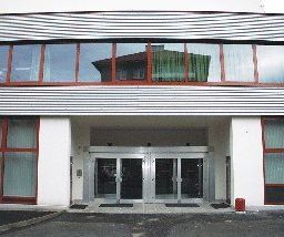 Ein modernes Gebäude mit Glasfenstern und Türen, einem großen Bogeneingang und einer reflektierenden Glasfassade. Der Eingang wird von rot umrahmten Fenstern flankiert, und in der Nähe befindet sich ein kleiner roter Feuerlöscher.