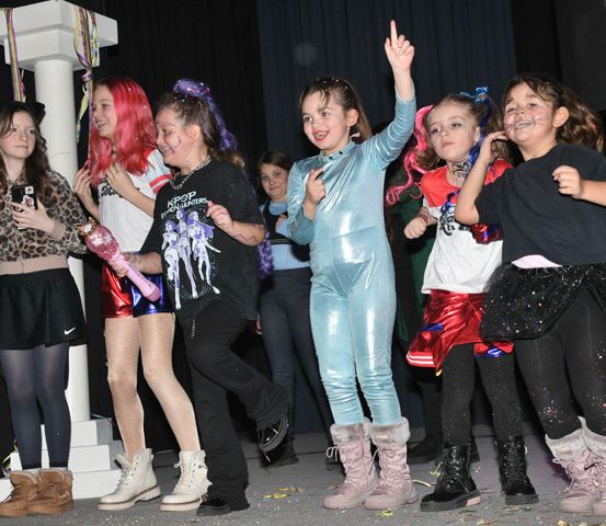 Eine Gruppe junger Mädchen in verschiedenen Kostümen tritt auf der Bühne auf, eine zeigt nach oben, während andere lächeln und Gegenstände halten. Links steht eine weiße Säule mit bunten Luftballons, rechts stehen Lautsprecher.