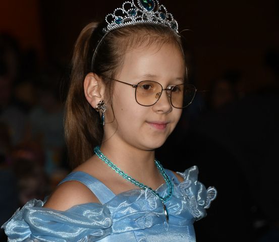 Ein junges Mädchen mit blauem Kleid und Krone, lächelt mit einer Brille und einer blauen Halskette. Der Hintergrund ist unscharf.