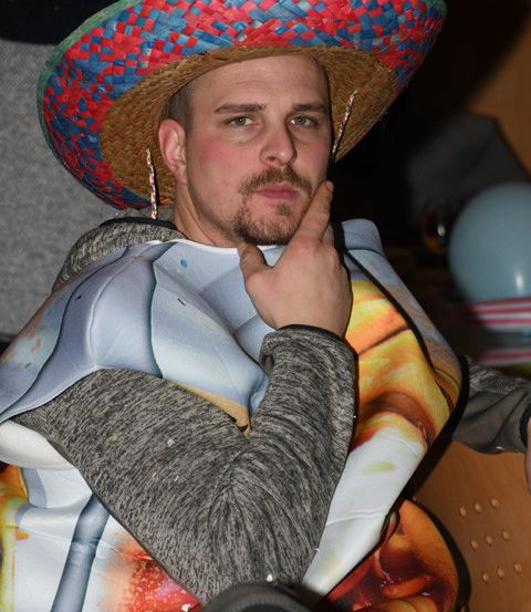 Ein Mann mit einem Sombrero und einem grauen Langarmshirt mit Tortilla-Aufdruck sitzt drinnen. Er hat die Hand am Kinn und schaut zur Seite. Im Hintergrund ist eine verschwommene Umgebung mit Luftballons und einem Tisch.