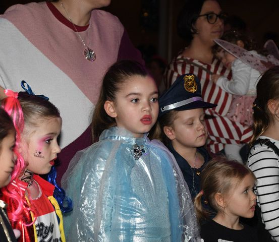 Eine Gruppe von Kindern in verschiedenen Kostümen, darunter eine Polizistin, stehen zusammen und schauen sich etwas an. Dahinter stehen Erwachsene, einer hält ein Kind mit Flügeln.