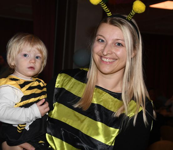 Eine Frau in einer Bienenkostüm, die ein kleines Kind in einem Bienenkostüm hält, beide lächeln bei einer Indoor-Veranstaltung.