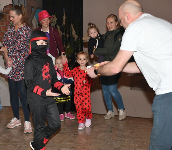 Eine Gruppe von Menschen tanzt in einem Raum. Ein Kind in einem Kostüm hält einen gelben Gegenstand. Ein Mann in einem weißen T-Shirt verteilt etwas. Die Frau im Hintergrund hält ein Baby. Im Hintergrund befindet sich eine Ziegelwand.