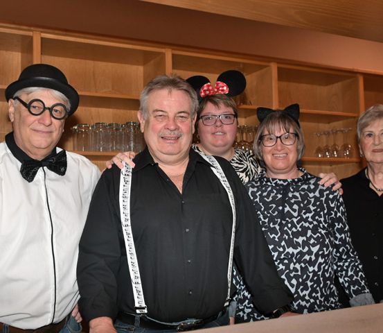 Fünf Erwachsene stehen eng zusammen für ein Foto. Sie tragen Kostüme, einschließlich Brillen, Hüte und Accessoires. Der Mann in der Mitte trägt Hosenträger. Dahinter befindet sich ein Holzregal mit Gläsern und eine Flasche obenauf.