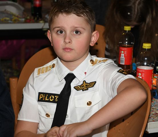 Ein junger Junge in einem Pilotenuniform sitzt an einem Tisch, die Hände ineinander verschränkt, mit anderen Personen im Hintergrund.