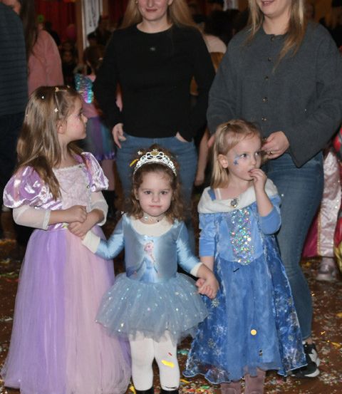 Drei junge Mädchen in Prinzessinenkostümen, die Händchen halten, eines mit einer Tiara, umgeben von Erwachsenen in einem festlichen, mit Konfetti gefüllten Raum.