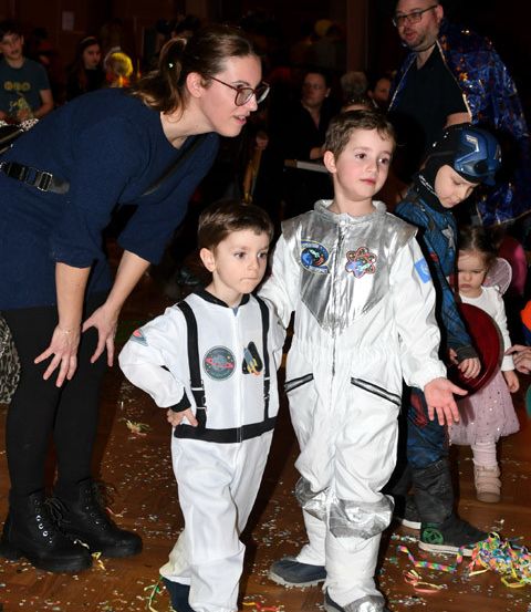 Eine Frau und zwei Kinder in Astronautenanzügen auf einer Party. Konfetti verstreut auf dem Boden. Andere Menschen in Kostümen im Hintergrund.