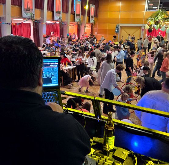 Ein DJ arbeitet auf einer Party, bei der eine große Menschenmenge tanzt, einige mit Luftballons. Tische und Stühle sind aufgestellt, und einige Leute sitzen.