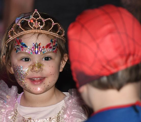 Ein kleines Mädchen mit glitzerndem Gesichtsschmuck und einer goldenen Krone lächelt und schaut einen Jungen in einem roten Hut an.
