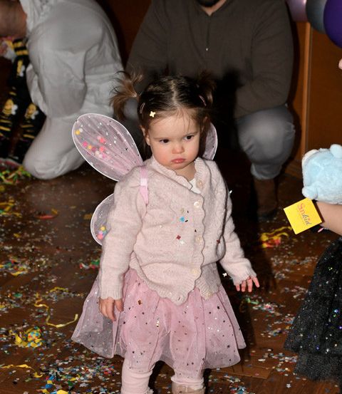 Ein kleines Mädchen in einem rosa Feenkostüm steht auf einem mit Konfetti bedeckten Boden und blickt nach links. Hinter ihr hockt ein Mann, und im Hintergrund steht eine Person in einem weißen Kostüm mit Luftballons.