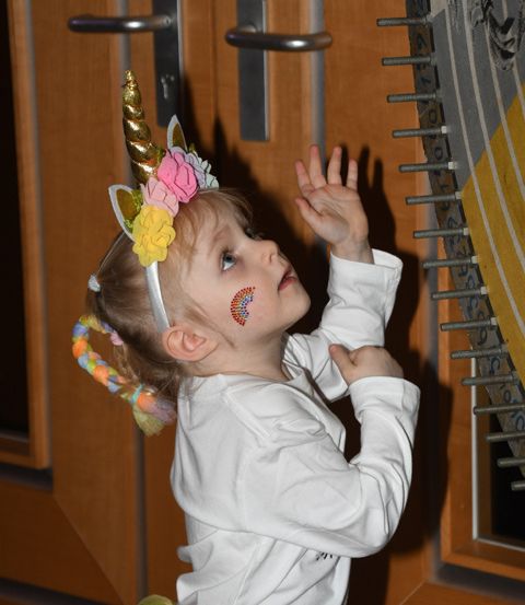 Ein kleines Mädchen in einem Einhornkostüm steht vor einer Holztür mit silbernem Griff und schaut mit erhobenen Händen nach oben.
