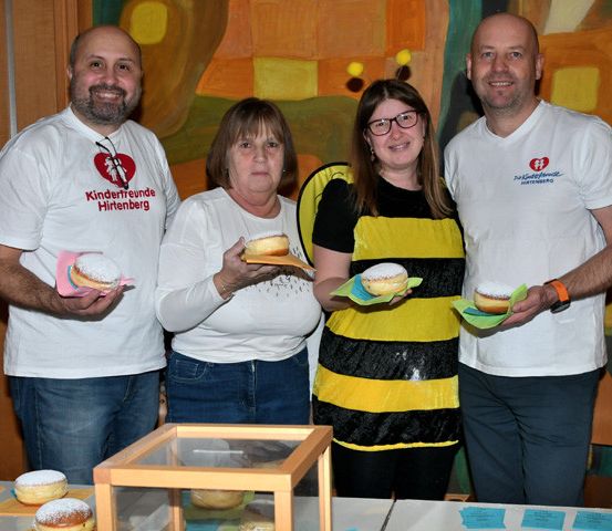 Vier Personen stehen lächelnd vor einem Tisch mit Donuts. Zwei Männer tragen weiße T-Shirts mit Logos, und eine Frau in einer Bienenkostüm hält einen Donut. Ein farbenfroher Hintergrund ist hinter ihnen.