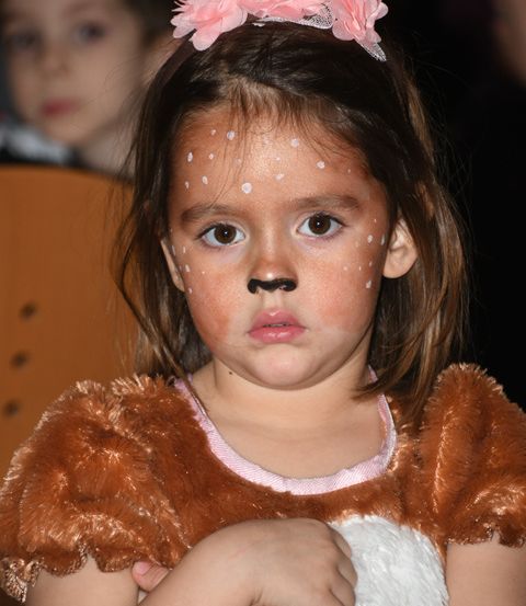 Ein kleines Mädchen in einem Hirschkostüm, mit weißen Punkten im Gesicht und einer rosa Blumenhaarband, sitzt mit verschränkten Armen.