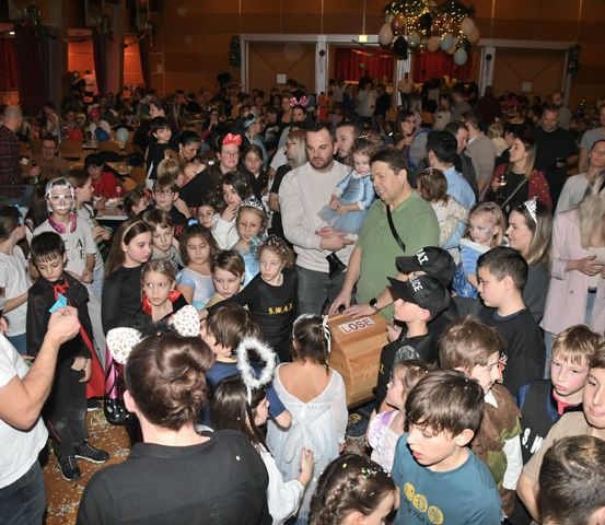 Eine Menschenmenge, darunter Kinder in Kostümen, versammelt sich um eine Holzkiste, möglicherweise für eine Tombola. Erwachsene sind anwesend, einer hält ein Baby. Der Raum ist für eine festliche Veranstaltung dekoriert.