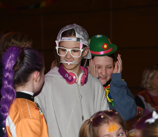 Eine Gruppe von Kindern, einige in Kostümen, stehen in einem Raum. Ein Mädchen mit lila Haaren und einer weißen Maske trägt eine graue Hoodie. Ein Mädchen mit einer grünen Mütze lächelt und ein anderes Mädchen berührt ihr Gesicht.
