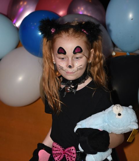 Ein junges Mädchen in einem Katzenkostüm mit Katzengesichtsbemalung, das ein blaues Plüschtier hält. Hinter ihr sind verschiedene bunte Luftballons zu sehen.