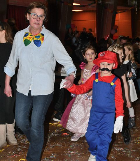 Eine Frau mit einer bunten Schleife geht mit einem als Mario verkleideten Kind durch einen überfüllten Raum mit Konfetti auf dem Boden.