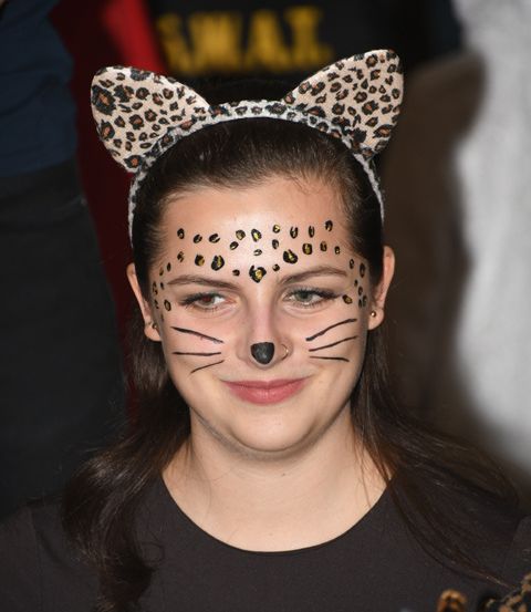 Ein junges Mädchen trägt eine Leopardenkopfbedeckung und hat ihr Gesicht in Katzen-Make-up gemalt, inklusive Schnurrhaaren und einer Nase. Sie lächelt.