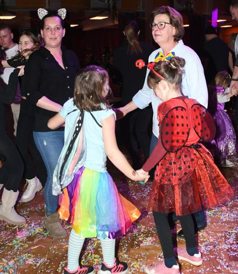 Eine Gruppe von Menschen tanzt auf einem hölzernen Boden mit Konfetti in einer festlichen Umgebung. Zwei junge Mädchen in bunten Kleidern halten die Hände einer Erwachsenen.