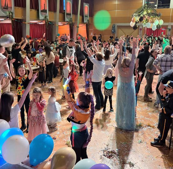 Eine große Gruppe von Menschen, darunter Kinder in Kostümen, tanzt in einem mit Ballons und festlichen Dekorationen gefüllten Raum. Einige heben die Hände, während andere Ballons halten. Der Raum ist mit Weihnachtsbäumen und Lichtern geschmückt.