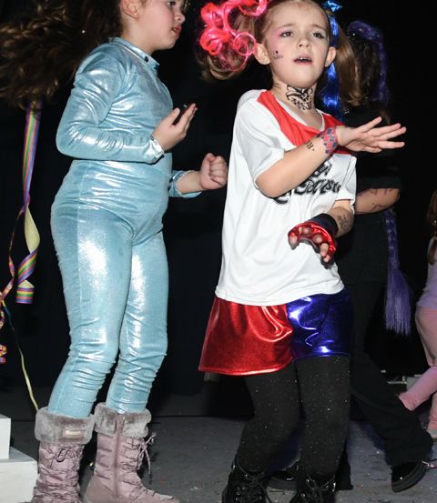 Zwei junge Mädchen in Kostümen stehen zusammen, eines in einem silbernen Jumpsuit, das andere in einem rot-blauen Kostüm mit weißem Shirt. Sie befinden sich wahrscheinlich auf einer Kostümparty.