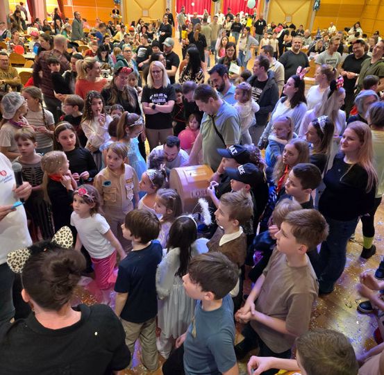 Eine große Menschenmenge versammelt sich drinnen, Erwachsene und Kinder gleichermaßen, einige in Kostümen, andere halten Luftballons. Ein Mann in Weiß hält ein Mikrofon. Der Raum ist mit Luftballons und Wimpelketten geschmückt.
