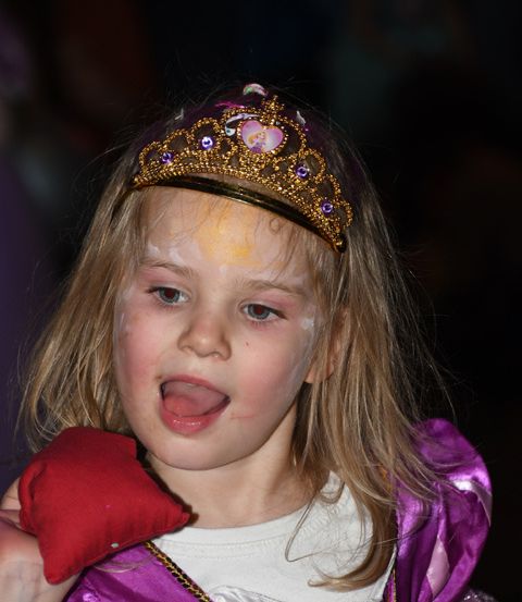Ein junges Mädchen in einem lila Kleid und einer goldenen Krone streckt die Zunge heraus und hält ein rotes Kissen.