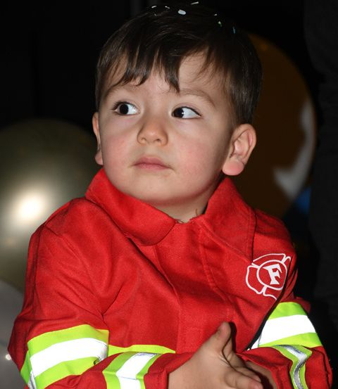 Ein kleiner Junge in einem roten Feuerwehrmann-Jacke sitzt mit gefalteten Händen vor sich und schaut zur Seite. Hinter ihm befinden sich zwei Luftballons, einer rot und einer weiß.
