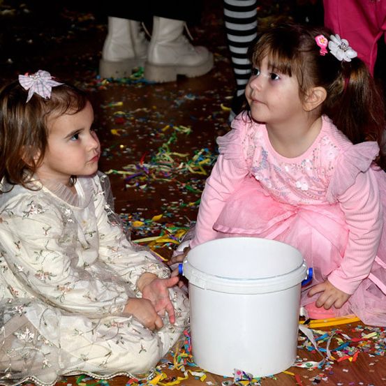 Zwei kleine Mädchen sitzen auf einem Boden mit Konfetti. Ein Mädchen hält einen Eimer, das andere sieht ihn an. Hinter ihnen steht eine Person in rosa Hosen.
