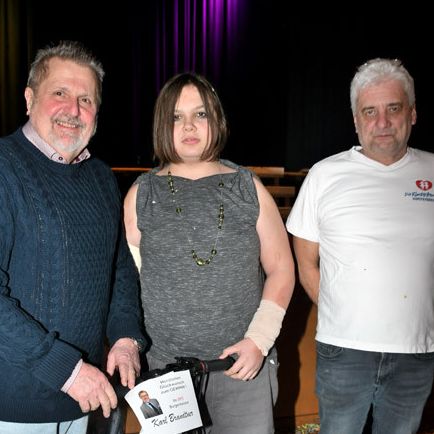 Drei Menschen stehen zusammen in einem Raum und halten einen Roller. Eine Frau mit einem bandagierten Arm und einem lächelnden Mann neben ihr. Ein Mann in einem weißen T-Shirt steht neben ihnen.