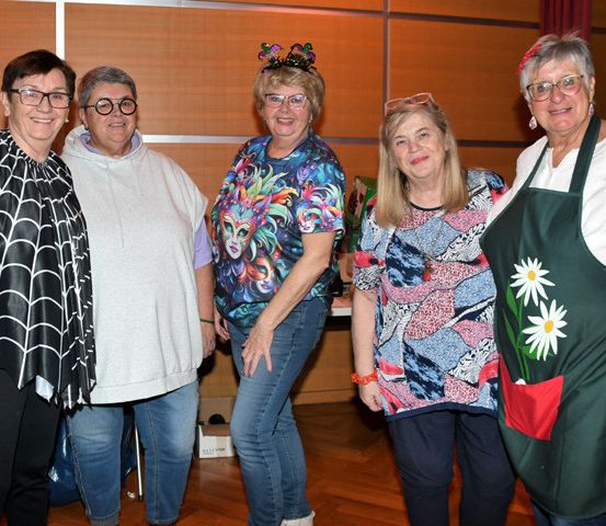 Fünf Frauen stehen lächelnd zusammen, mit einer hölzernen Wand und roten Vorhängen im Hintergrund.
