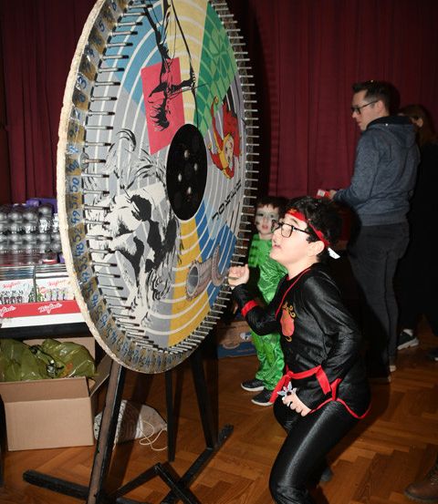 Ein Kind in einem Kostüm wirft ein Objekt auf eine große Dartscheibe mit verschiedenen Bildern.