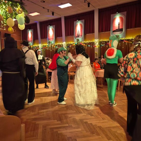 Mehrere Menschen in Kostümen tanzen in einem dekorierten Raum. Eine Person ist als Nonne verkleidet.