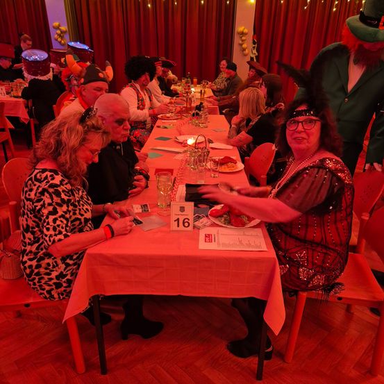 Eine Gruppe von Menschen sitzt an einem langen Tisch in einem Speisesaal. Eine Frau vorne hält einen Teller mit Essen. Der Tisch ist mit einer rosa Tischdecke bedeckt.