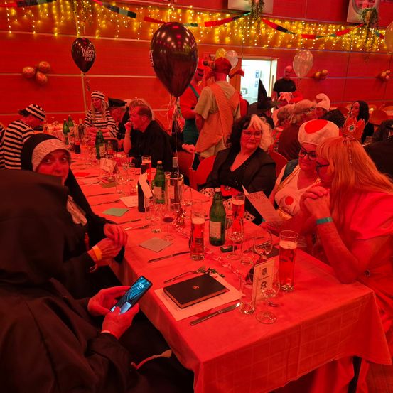 Eine Gruppe von Menschen in verschiedenen Kostümen sitzt an einem langen Tisch mit Getränken und Essen in einem dekorierten Raum. Einige tragen Hüte, Brillen und Armbänder. Über ihnen befinden sich Ballons und Lichter.