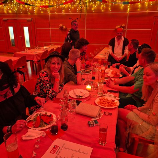 Eine Gruppe von Menschen sitzt an einem Tisch in einem Restaurant. Sie sind in verschiedenen Kostümen gekleidet, darunter Hüte und Masken. Der Tisch ist mit Tellern, Getränken und Kerzen gedeckt. Der Raum ist schwach beleuchtet mit roten Lichtern und mit hängenden Lichtern dekoriert.