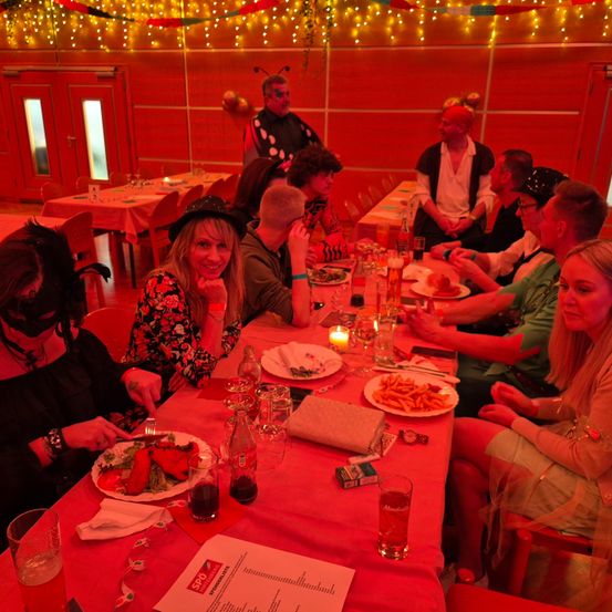 Eine Gruppe von Menschen in Kostümen sitzt an einem Tisch mit Essen, Getränken und einer Speisekarte. Sie scheinen eine Dinnerparty zu veranstalten, mit Dekorationen und einer festlichen Atmosphäre.