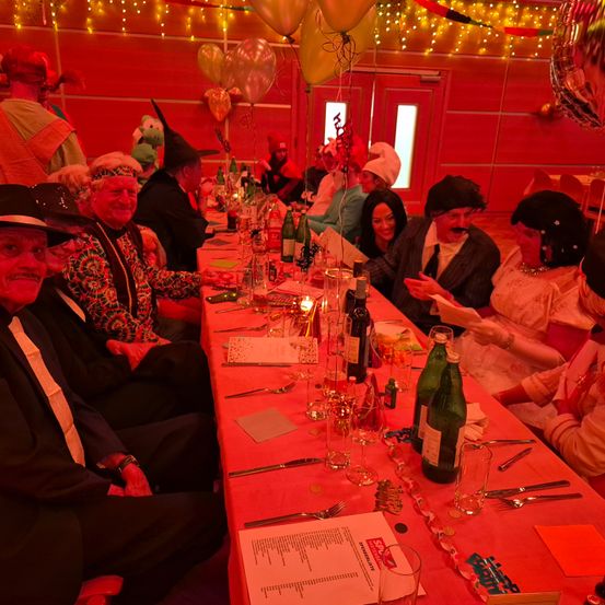 Eine Gruppe von Menschen sitzt an einem langen Tisch mit verschiedenen Gegenständen darauf. Der Tisch ist mit Flaschen, Gläsern und anderen Gegenständen gedeckt. Einige Leute tragen Hüte und andere Halsketten. Es gibt Luftballons im Raum.