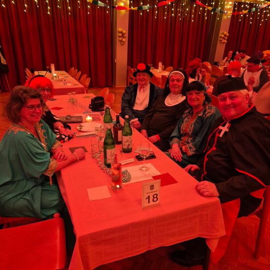 Eine Gruppe von Menschen sitzt an einem Tisch mit Weingläsern, Flaschen und der Tischnummer 18. Sie sind in festlicher Kleidung gekleidet. Der Tisch ist mit einer Tischdecke bedeckt. Der Raum ist mit roten Vorhängen und Luftballons dekoriert. Auf dem Tisch steht eine Kerze.