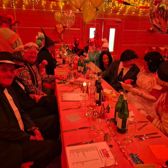 Eine Gruppe von Menschen in Kostümen sitzt an einem langen Tisch in einem Raum mit roten Lichtern und Dekorationen. Sie lächeln und scheinen sich zu amüsieren. Flaschen, Gläser und Teller stehen auf dem Tisch.