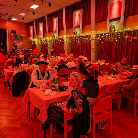 Ein festlicher Innenbereich mit roter Beleuchtung und Dekorationen. Viele Menschen sitzen an Tischen und tragen festliche Kostüme. Flaschen, Gläser und Teller sind auf den Tischen.